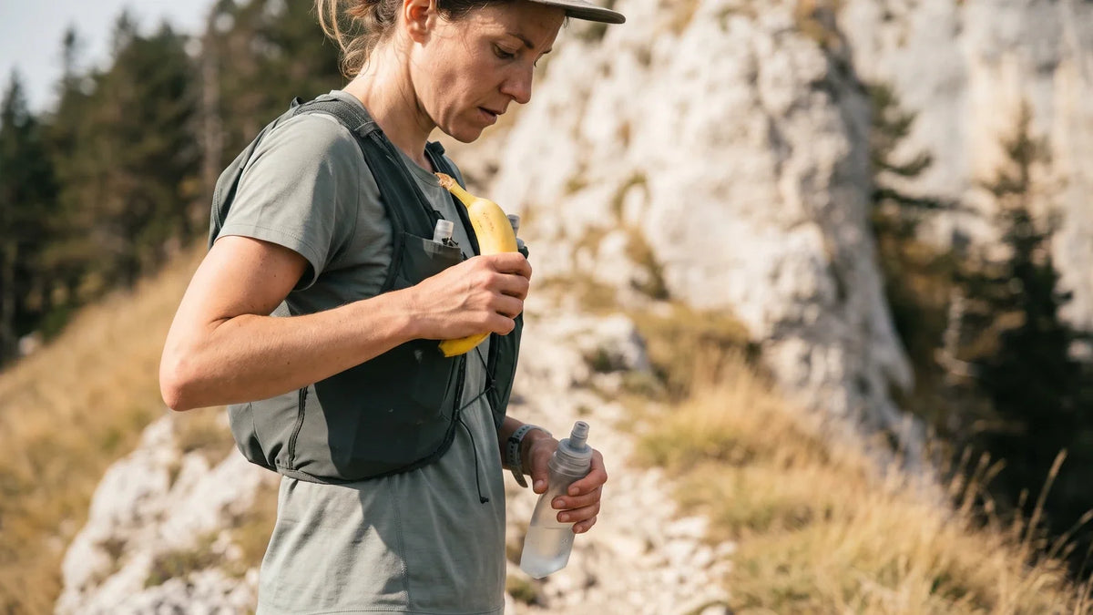 Coureuse de trail arrêtée sur un sentier au pied d'une falaise calcaire, une banane dans une main et une flasque souple dans l'autre