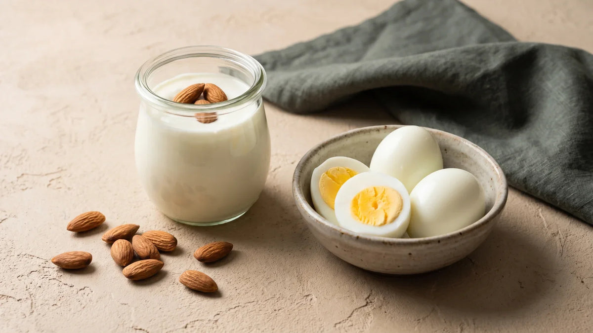 Un pot en verre de yaourt garni de trois amandes, un bol en grès contenant des œufs durs coupés en deux et des amandes éparpillées sur un plan de travail clair.
